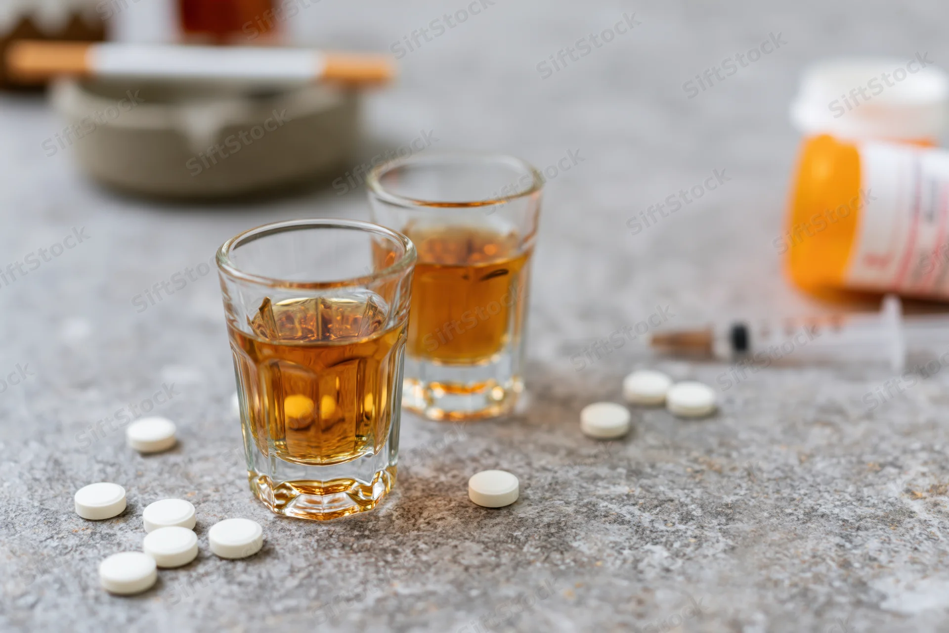 Two shot glasses filled with amber whiskey on a grey stone surface surrounded by scattered white pills, a syringe, a cigarette in an ashtray, and a prescription bottle