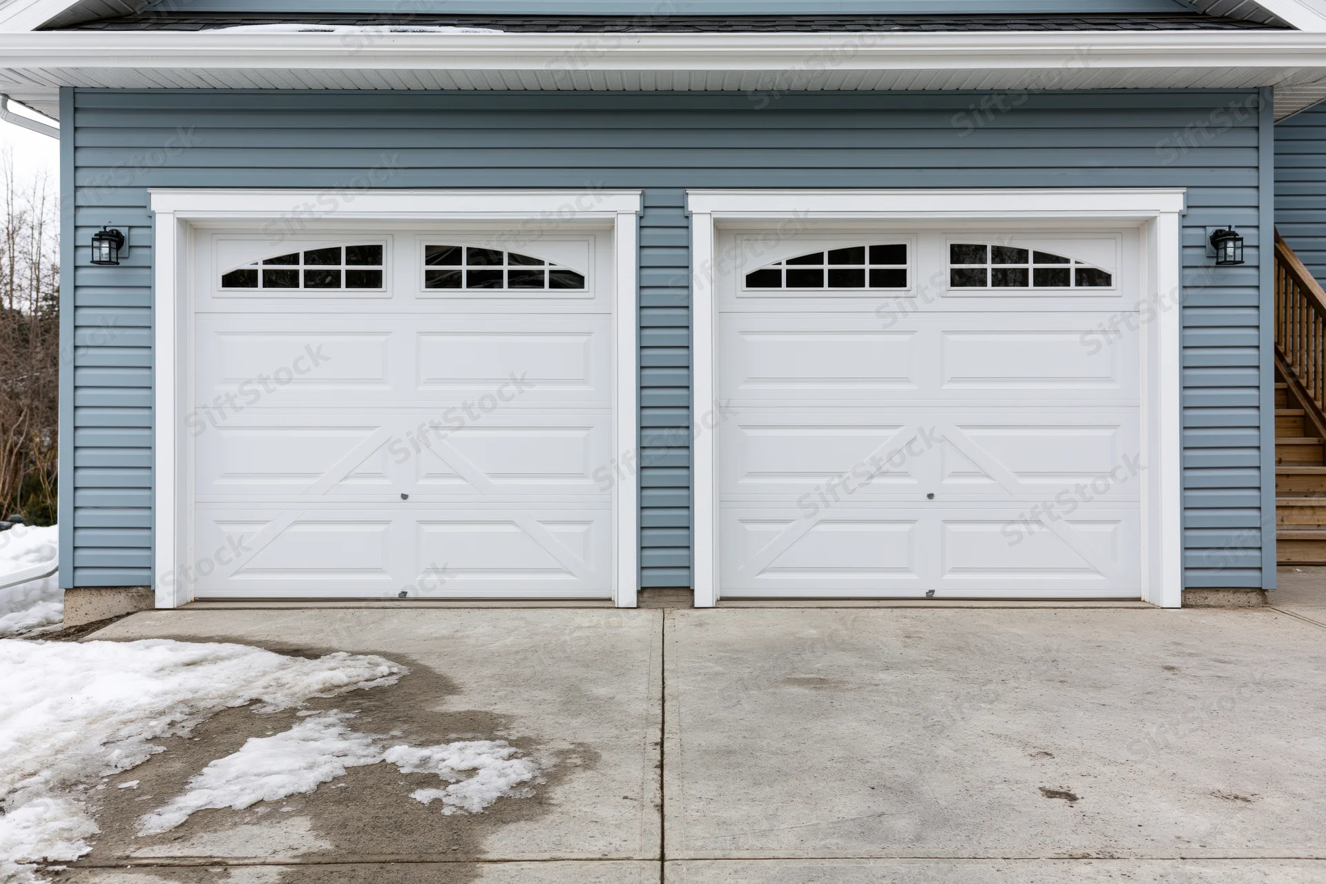 Two white raised-panel garage doors with arched transom windows on a blue-sided house exterior in winter