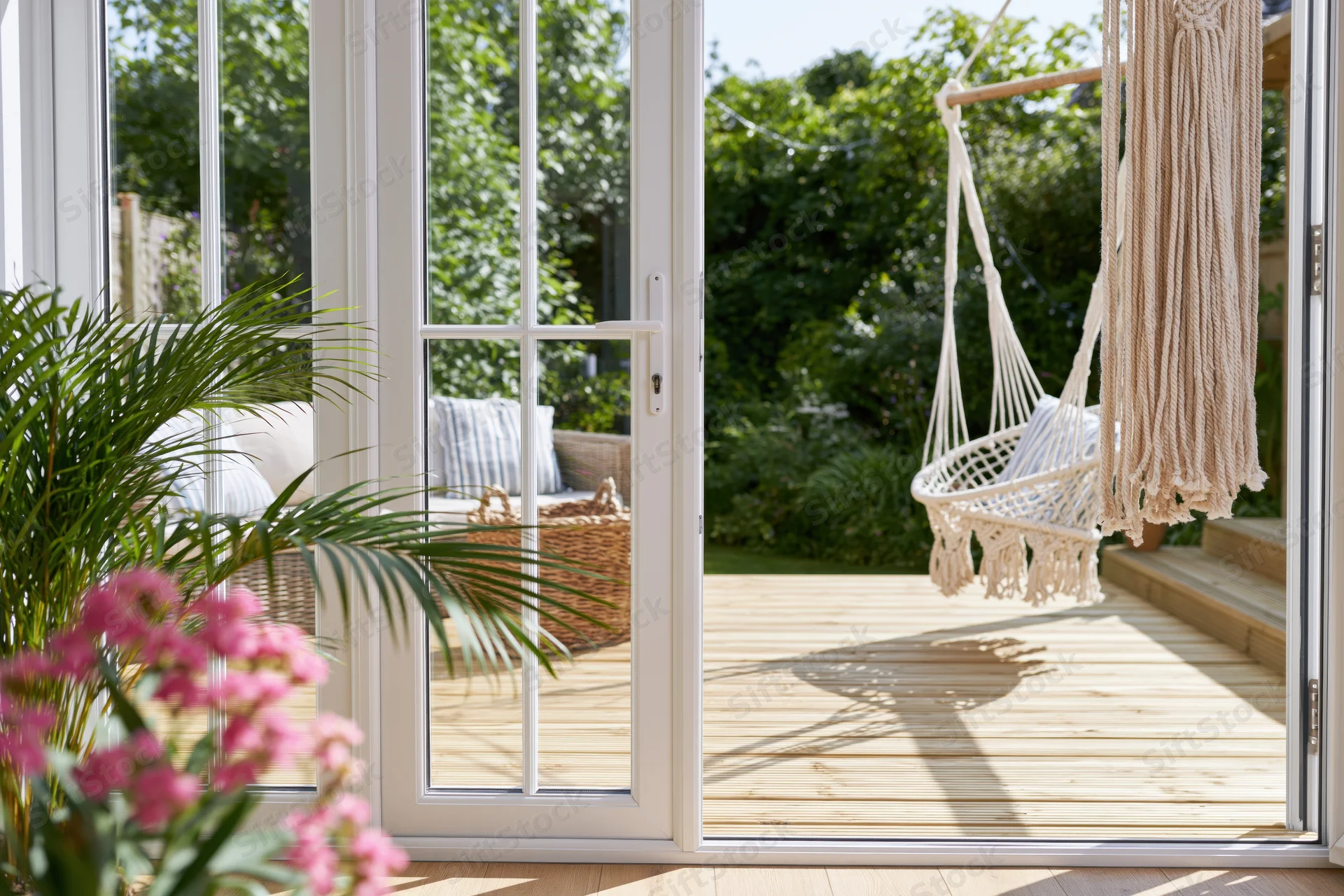 View through white-framed glass doors onto a sunlit wooden deck with a hanging macrame swing and garden greenery