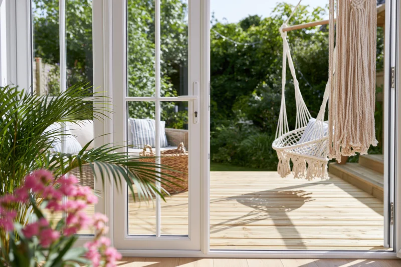 View through white-framed glass doors onto a sunlit wooden deck with a hanging macrame swing and garden greenery