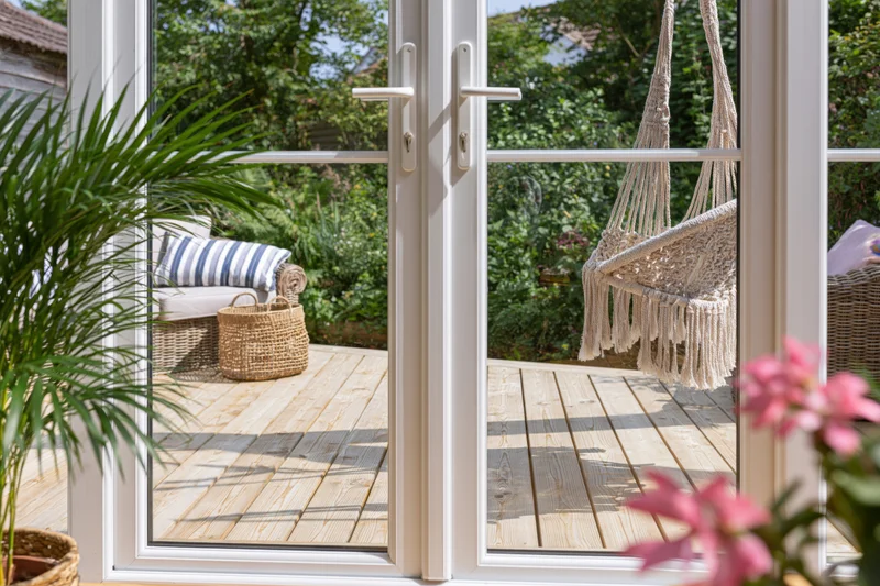View through white-framed glass patio doors onto a wooden deck with a macrame swing and garden greenery