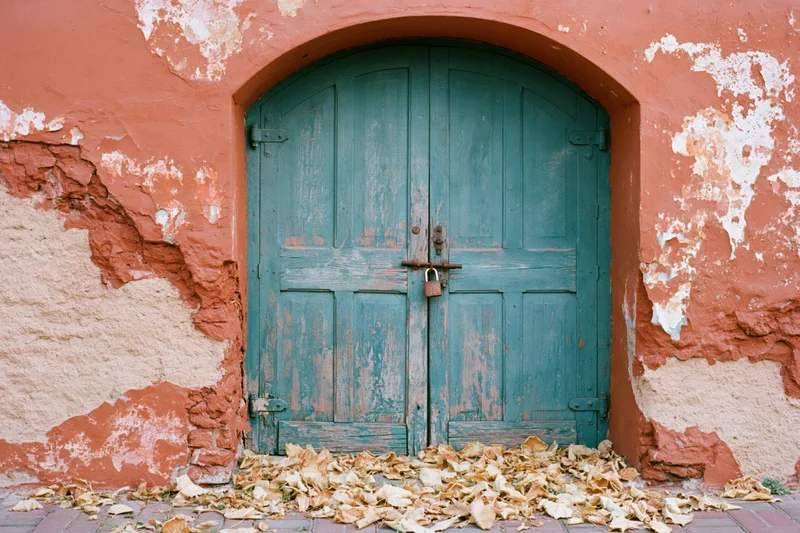 Weathered teal wooden double door in arched alcove with peeling orange stucco wall and fallen leaves on pavement