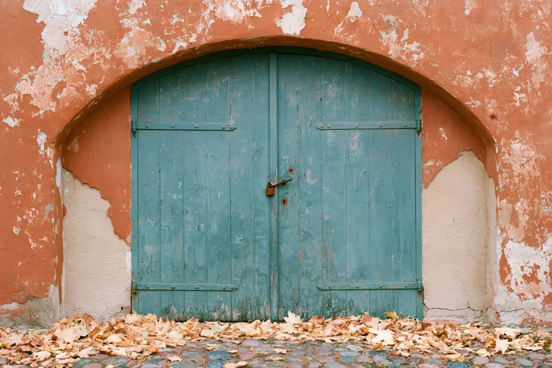Weathered teal wooden double door in arched alcove with peeling orange stucco and fallen leaves on cobblestone ground