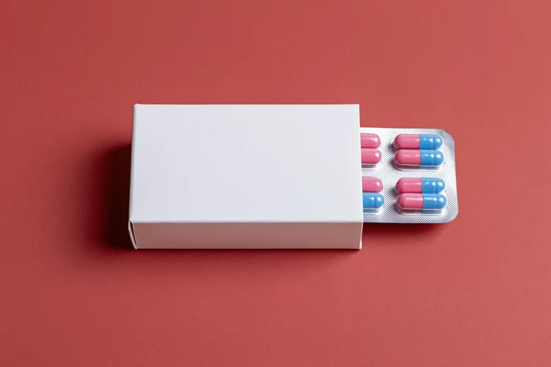 White medicine box partially open revealing pink and blue capsules in blister pack on red surface