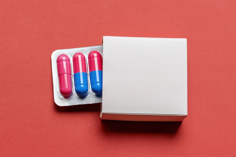 White medicine box partially open revealing blister pack of pink and blue capsules on red background