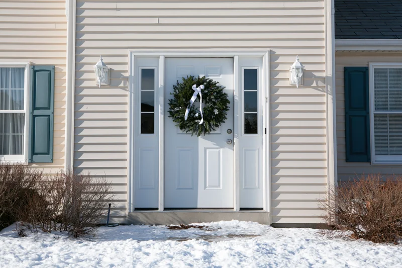 White front door with green christmas wreath and white bow on snowy porch of house with beige siding and blue shutters