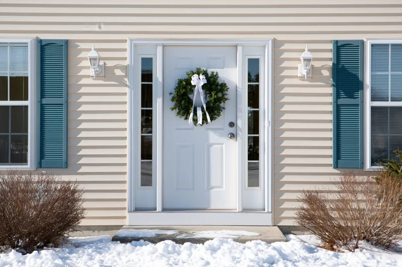 White front door with green christmas wreath on beige house exterior in snowy winter setting