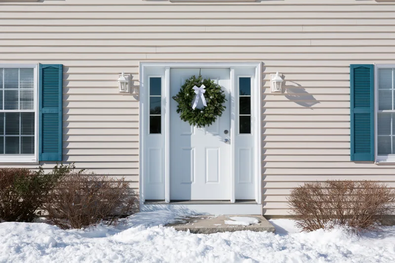 White front door with green christmas wreath on snowy porch of suburban house