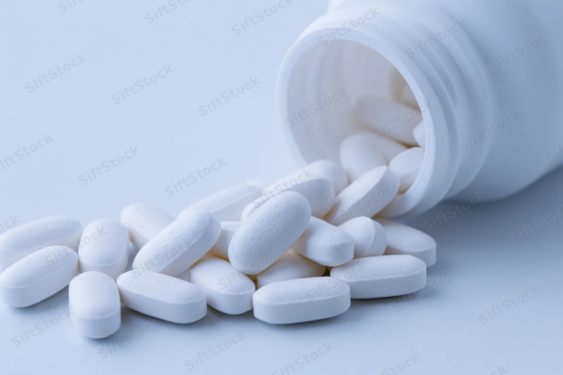 White oval pills spilling from an overturned plastic bottle on a light surface