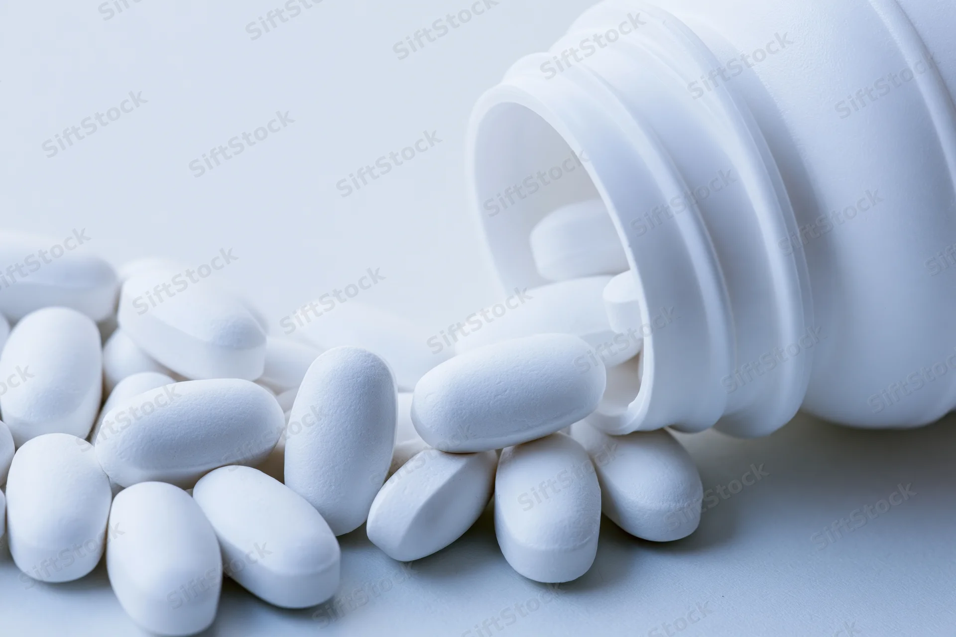White oval pills spilling from an overturned plastic bottle on a plain surface