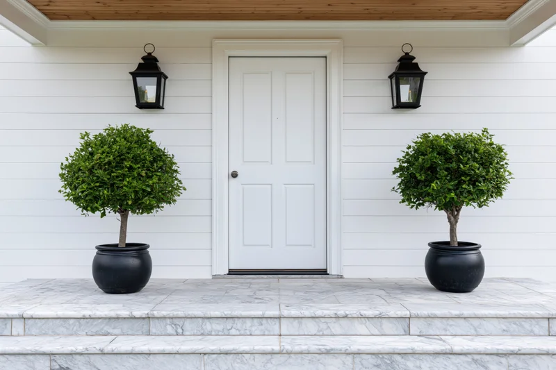 White front door with panel molding flanked by two potted topiary plants on marble steps under wooden ceiling with black lanterns