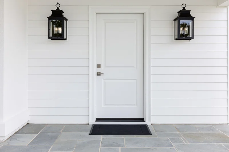 White raised-panel front door centered on a house exterior with black lantern lights and stone flooring