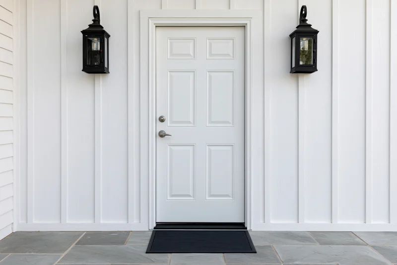 White raised-panel front door centered between black lanterns on a vinyl-siding house exterior