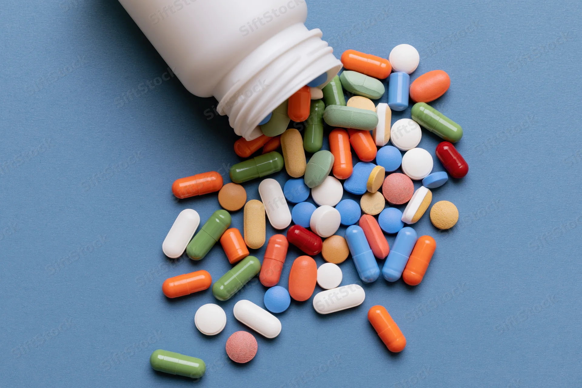 Assorted pills spilling from a white plastic bottle onto a blue surface