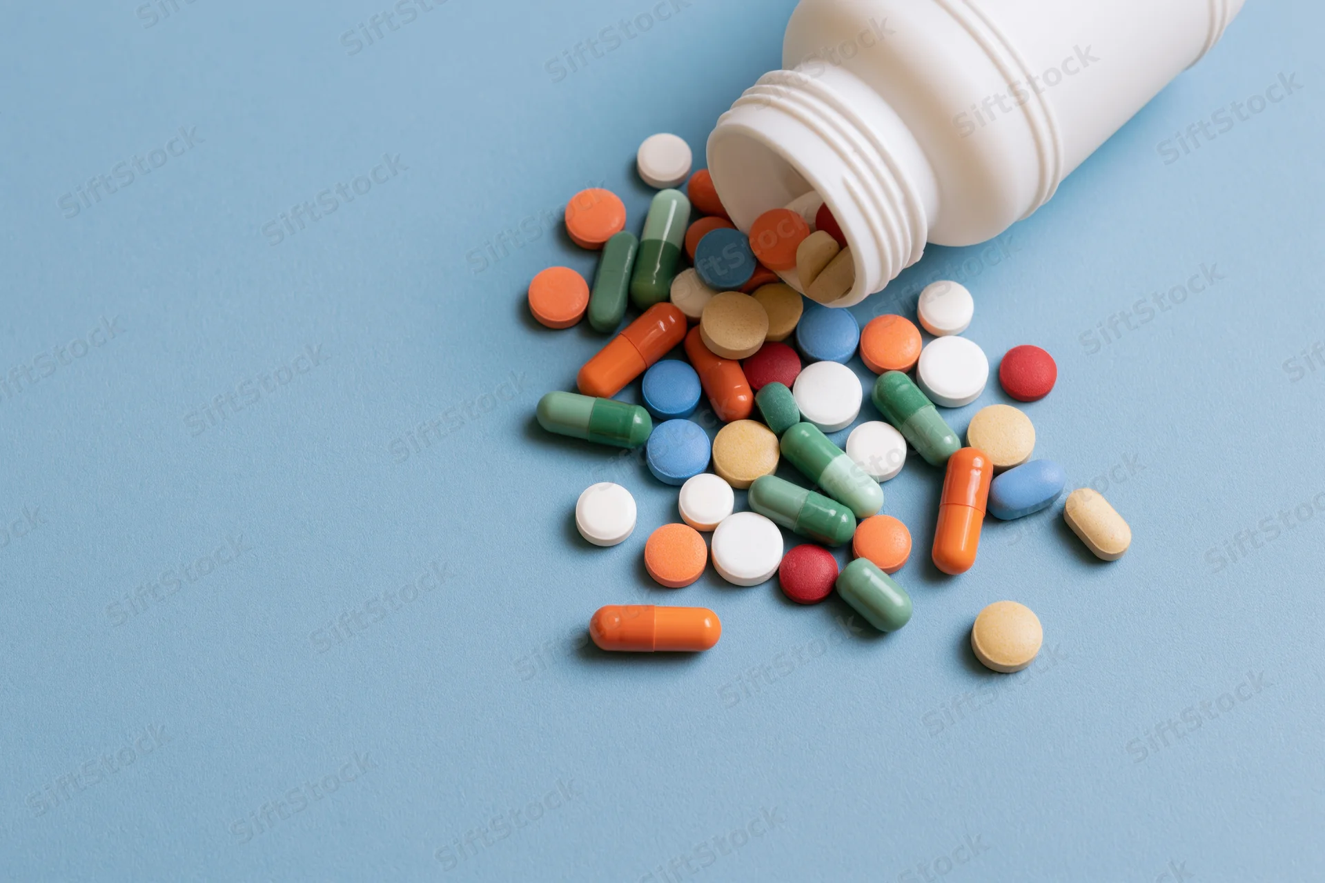Assorted pills and capsules spilling from a white plastic bottle onto a blue surface