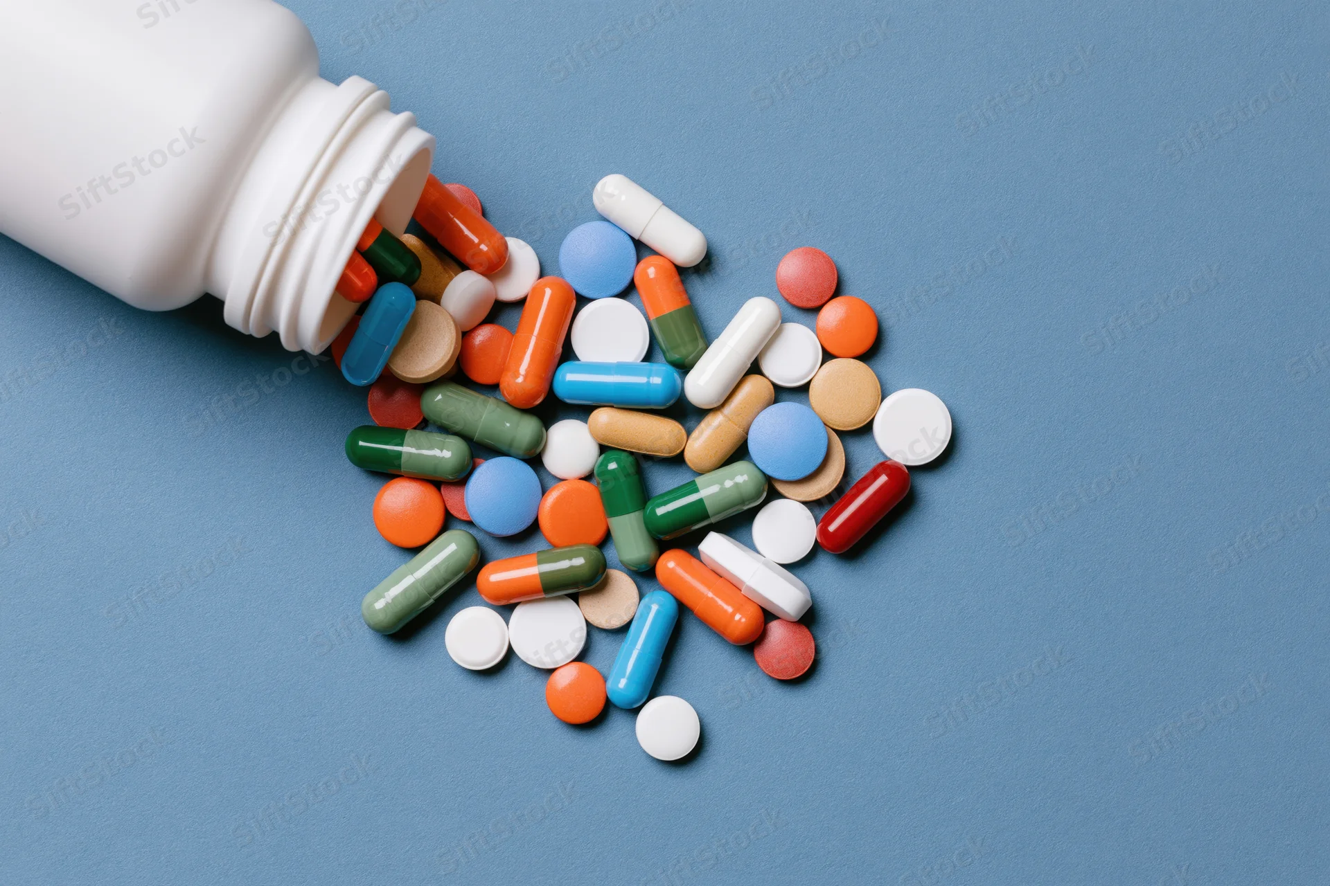 White plastic pill bottle lying on its side with assorted colorful pills spilling onto blue surface