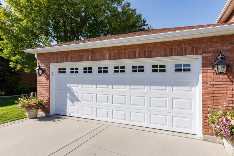 White raised-panel double garage door on brick house exterior with flower pots and driveway