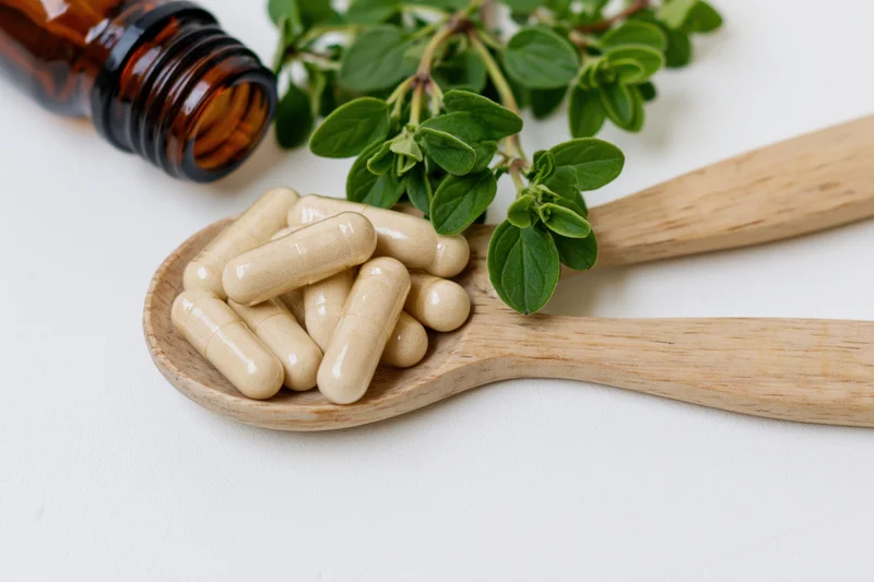 Wooden spoon holding herbal supplement capsules with fresh oregano and amber bottle on white surface