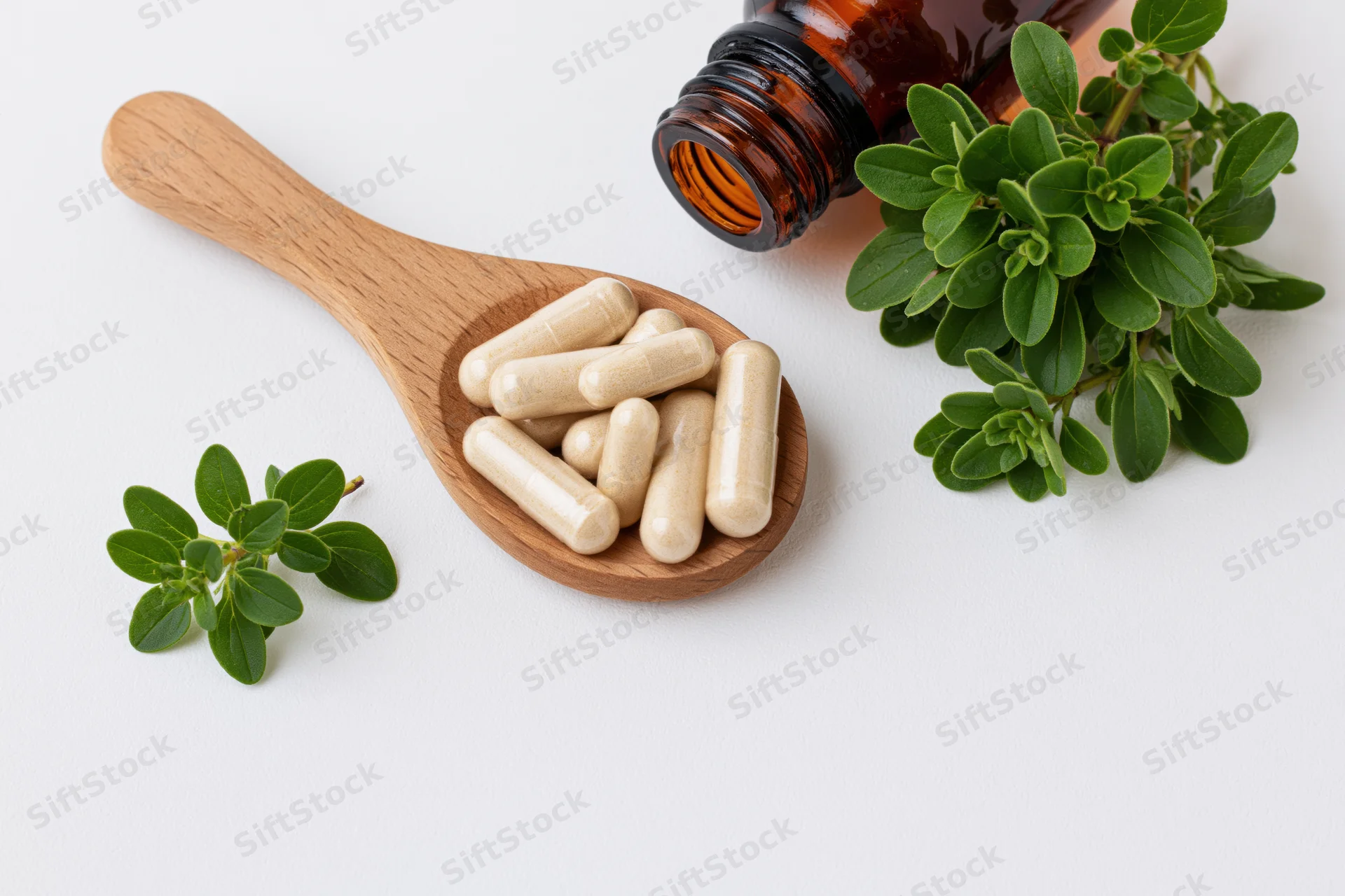 Wooden spoon holding herbal supplement capsules with fresh oregano leaves and amber bottle on white background