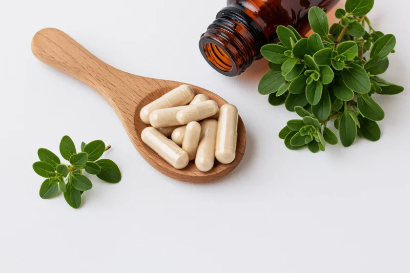Wooden spoon holding herbal supplement capsules with fresh oregano leaves and amber bottle on white background