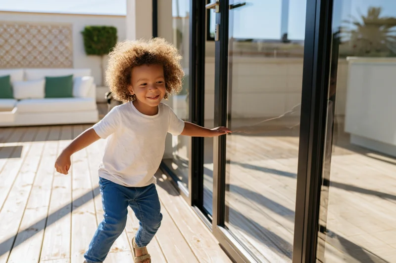 Young child with curly hair smiling while sliding open a glass patio door onto a wooden deck