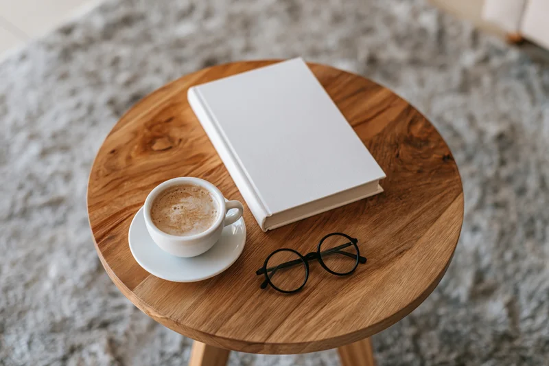 Blank white hardcover book with coffee cup and glasses on round wooden table over soft rug