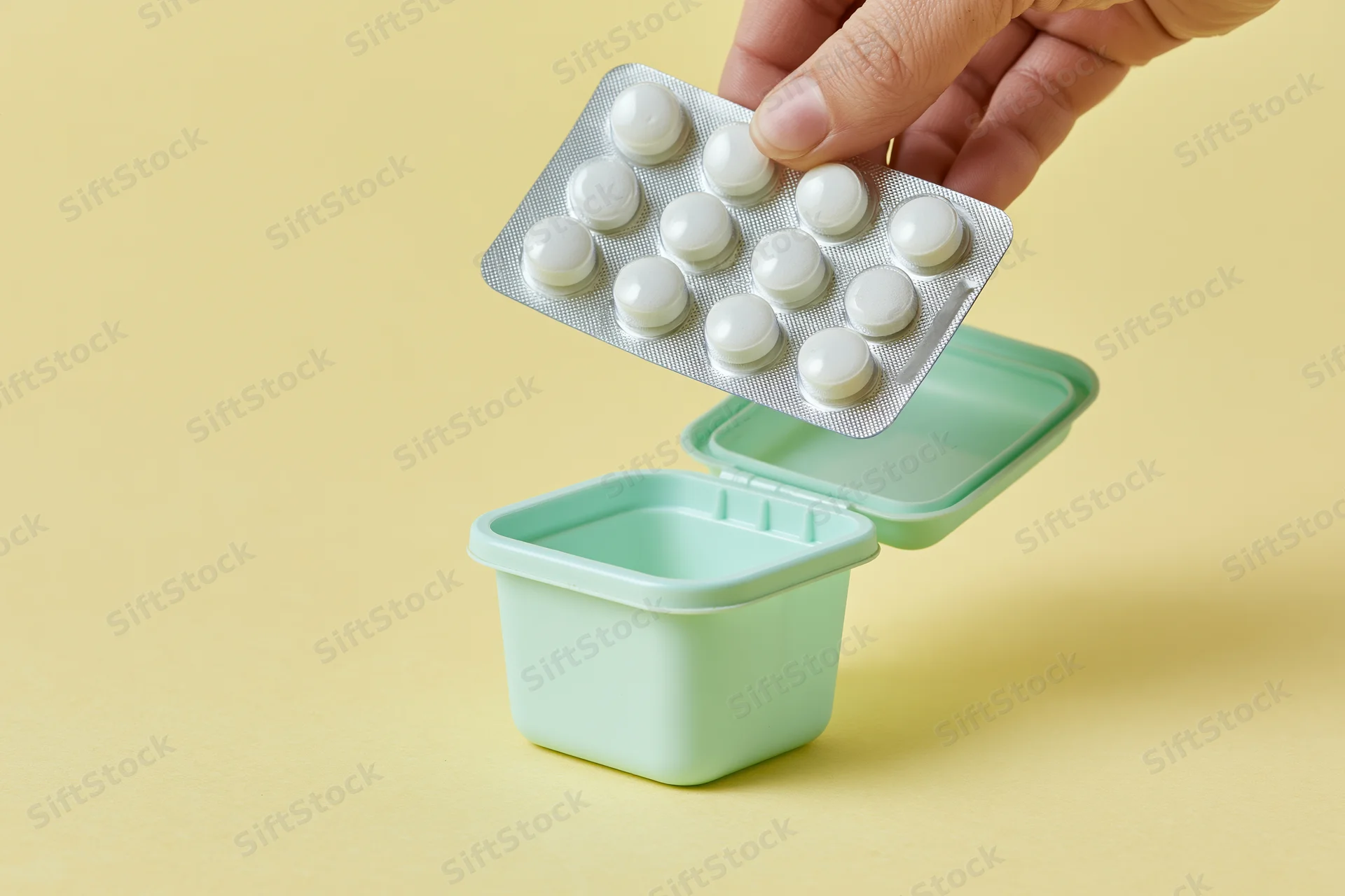 Hand dropping blister pack of white pills into small mint green container on yellow surface