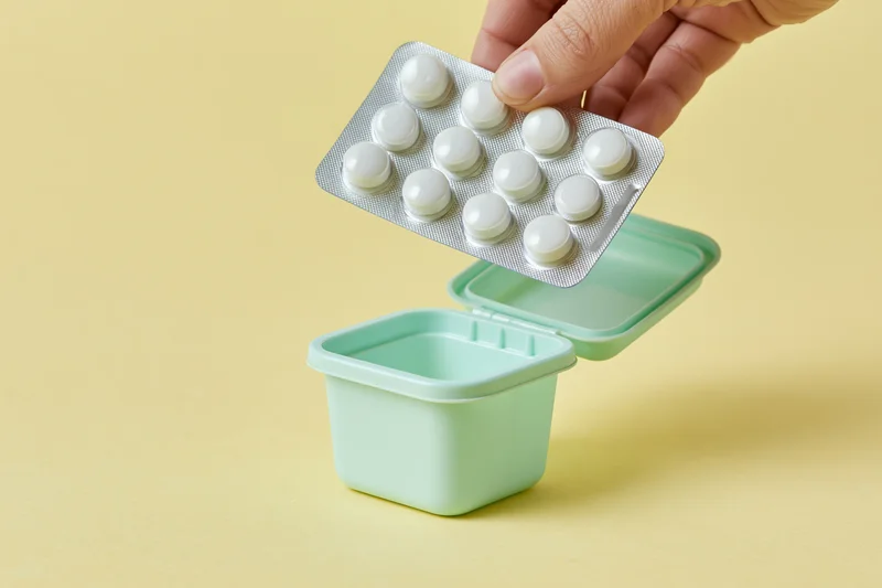 Hand dropping blister pack of white pills into small mint green container on yellow surface