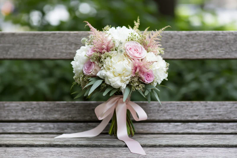 Bridal bouquet of white peonies and pink roses with astilbe and ribbon on a wooden bench in a garden