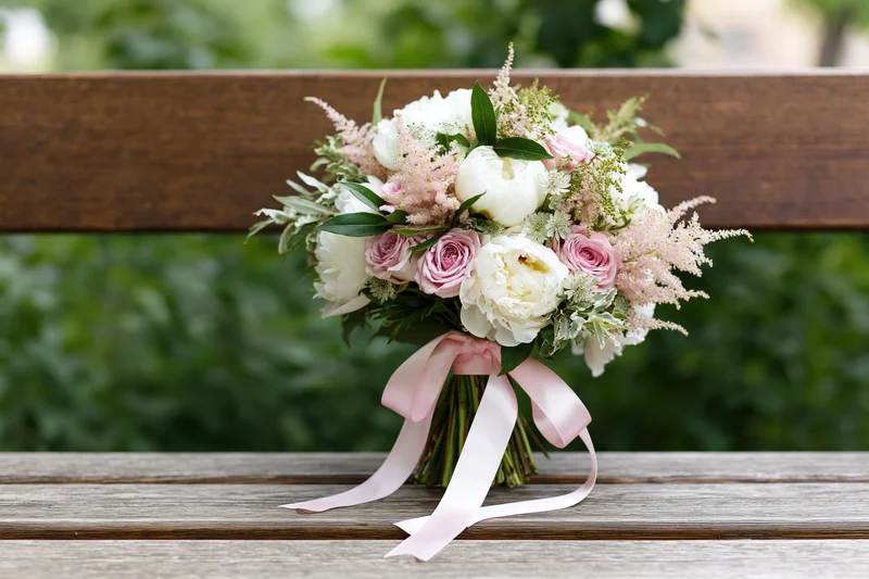 Bridal bouquet of white peonies, pink roses and astilbe resting on a wooden bench outdoors
