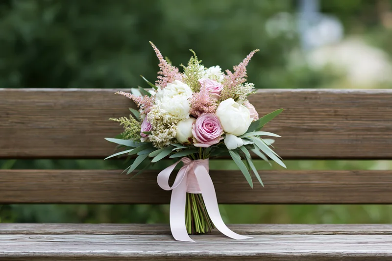 Bridal bouquet with white peonies, pink roses and astilbe resting on a wooden bench outdoors