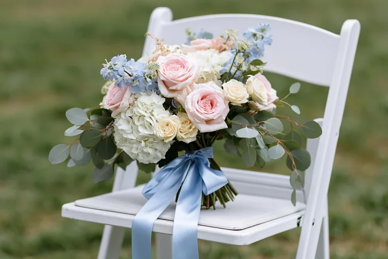 Bridal bouquet with pink roses, blue hydrangea and eucalyptus tied with satin ribbon resting on white folding chair outdoors