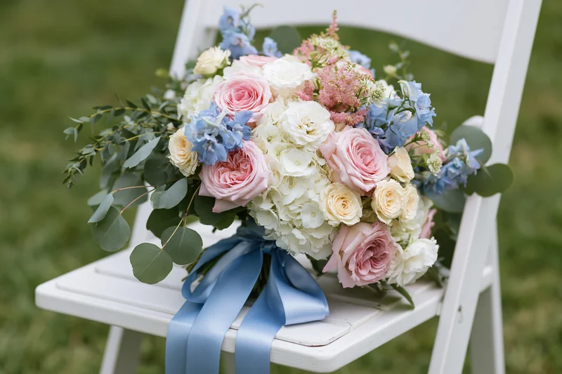 Bridal bouquet with pink roses, white hydrangeas, blue flowers and greenery tied with blue ribbon resting on white folding chair outdoors on grass