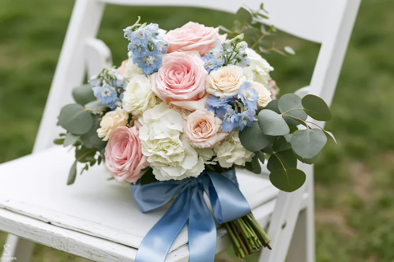 Bridal bouquet with pink roses, blue flowers and greenery tied with blue ribbon resting on white wooden folding chair outdoors