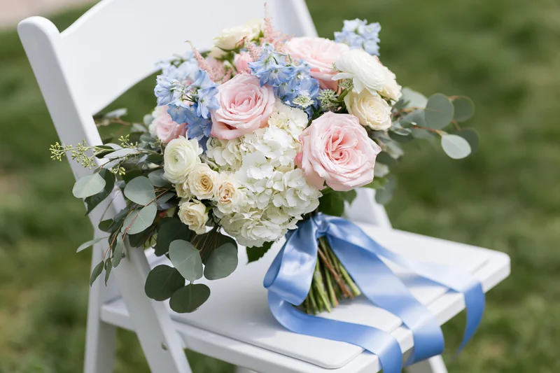Bridal bouquet with pink roses, blue flowers and greenery resting on white folding chair outdoors