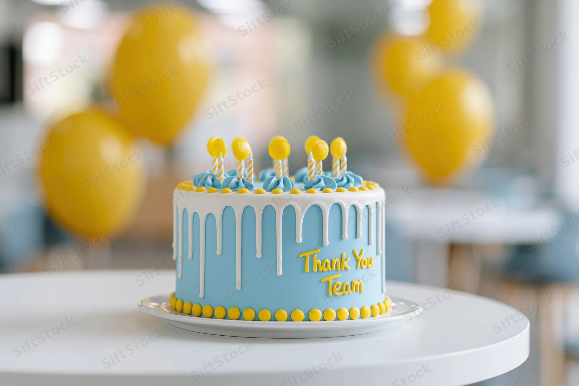 Blue and white celebration cake with yellow decorations and thank you team message on a table with balloons in the background