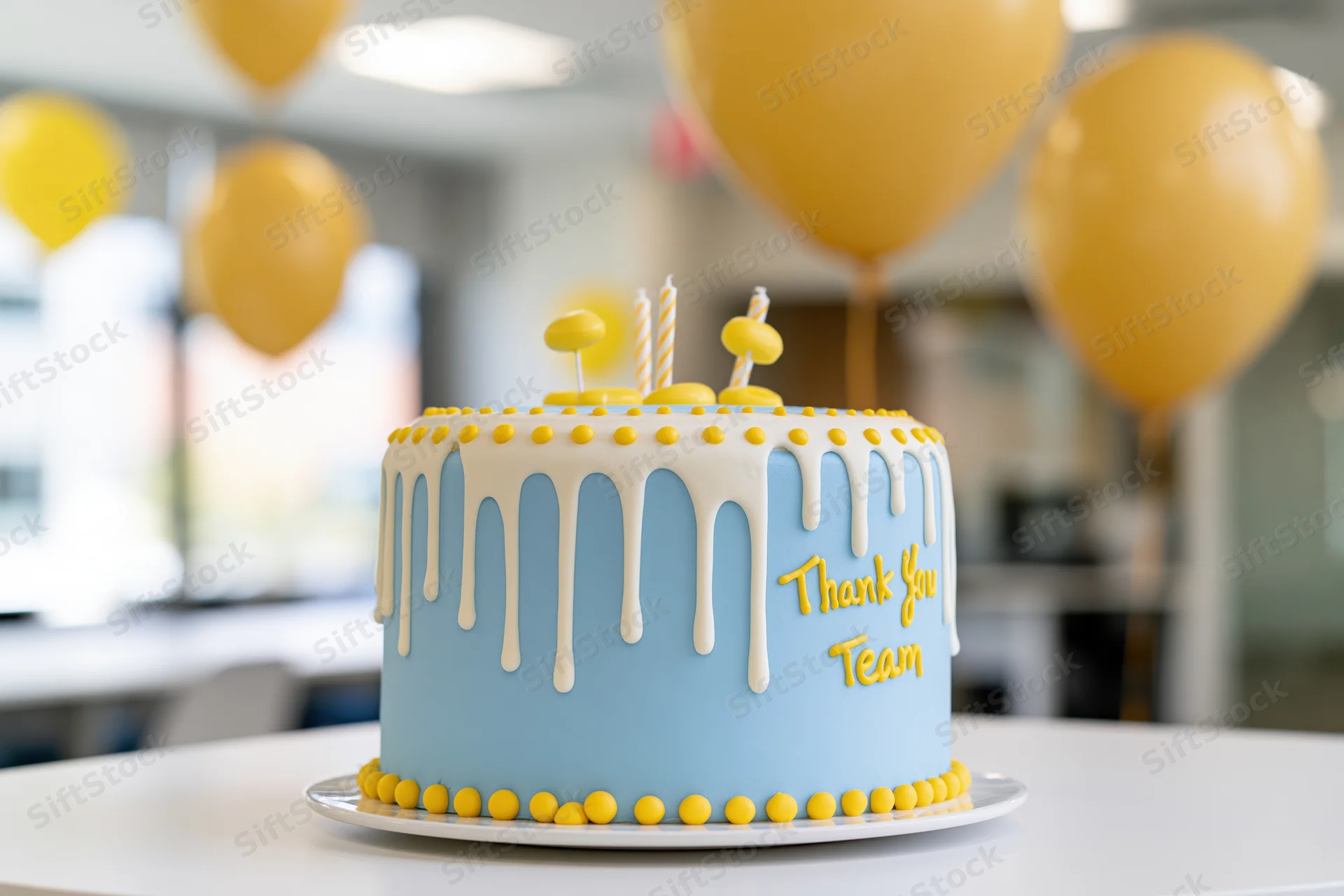 Blue and yellow fondant cake with white drizzle icing and thank you team message on a table in an office with balloons in the background