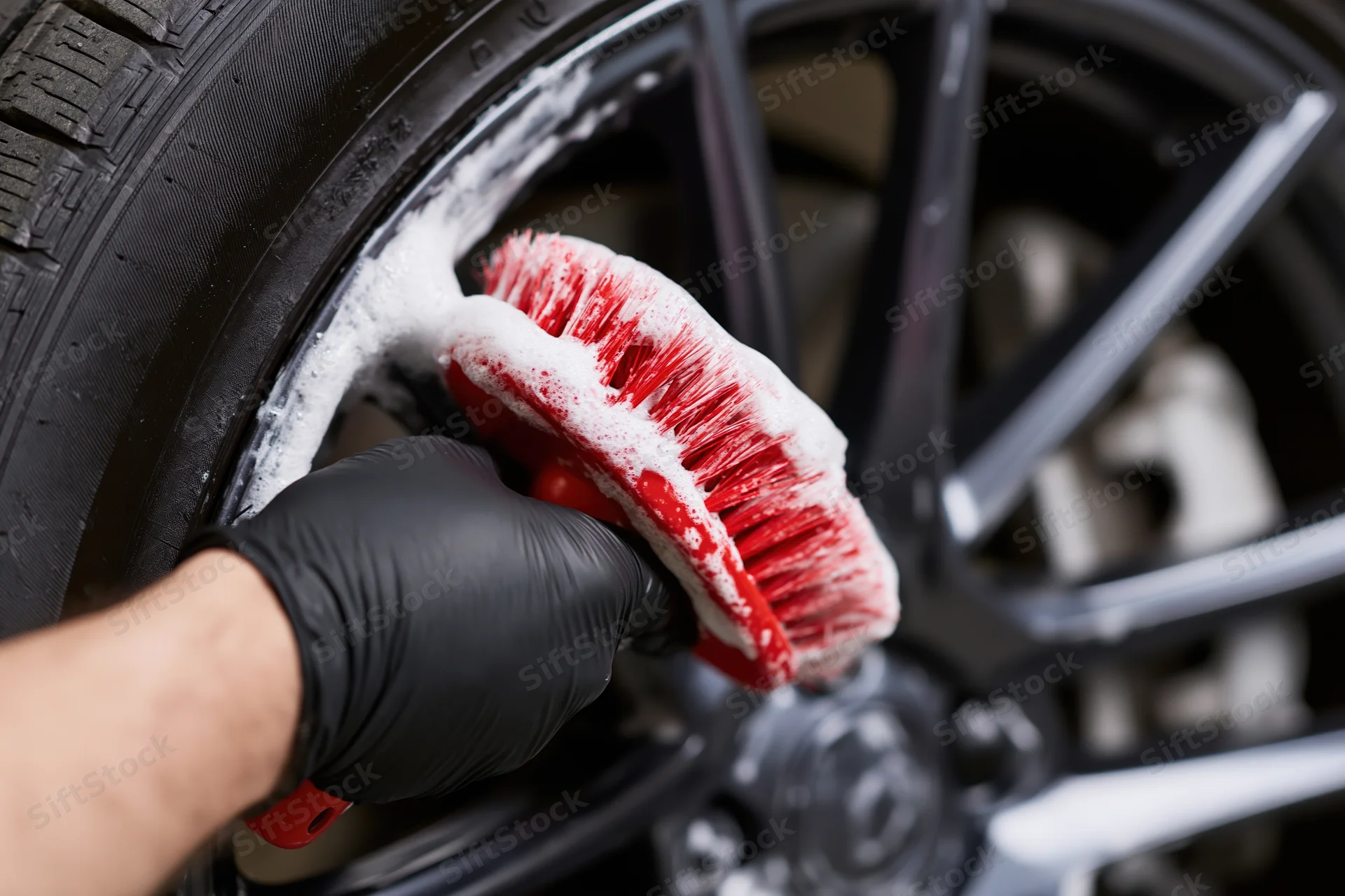 Hand in black glove scrubbing car wheel with red brush and white foam