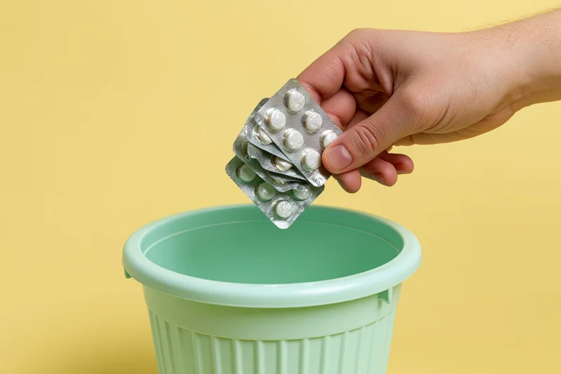 Hand dropping blister packs of white pills into a green trash bin