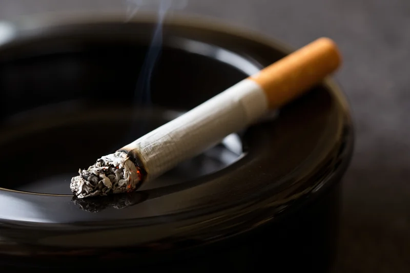 Lit cigarette with ash and smoke resting diagonally on a glossy black ashtray