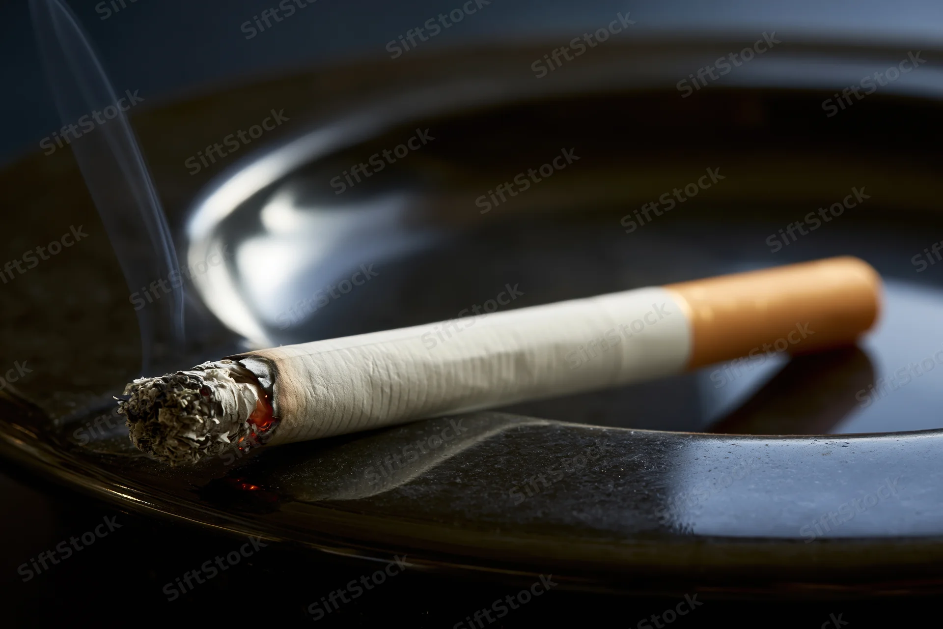 Lit cigarette with ash and smoke resting diagonally on a dark glossy black ashtray