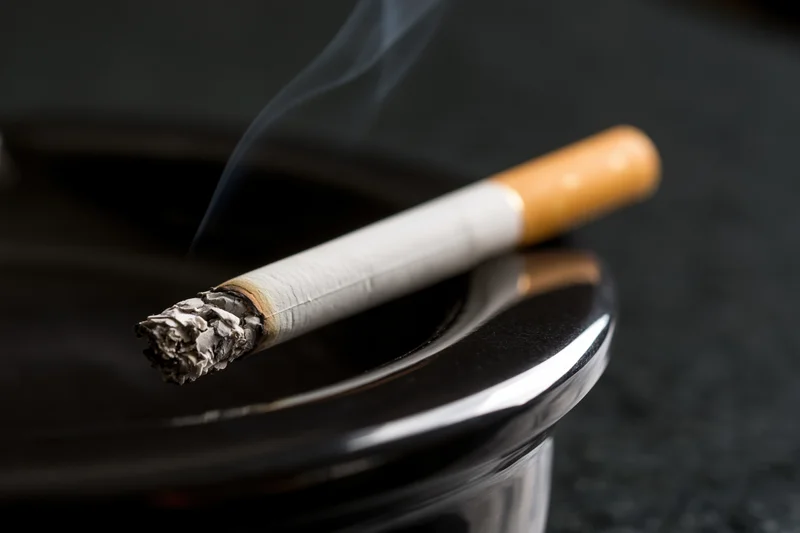 Lit cigarette with ash and smoke resting diagonally on a black glossy ashtray