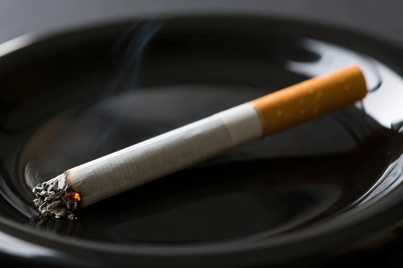 Lit cigarette with ash and smoke resting diagonally on a glossy black ashtray