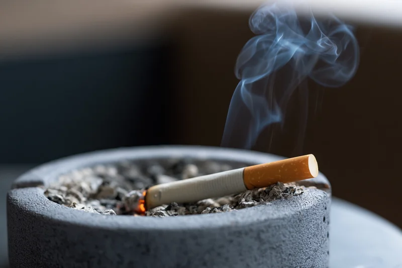 Lit cigarette burning in an ash-filled ceramic ashtray with rising smoke indoors