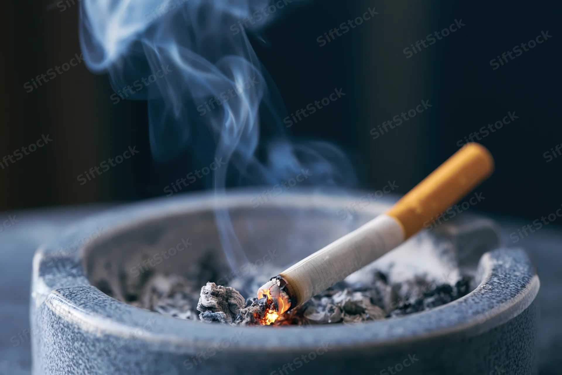Lit cigarette burning in an ash-filled ceramic ashtray with rising smoke