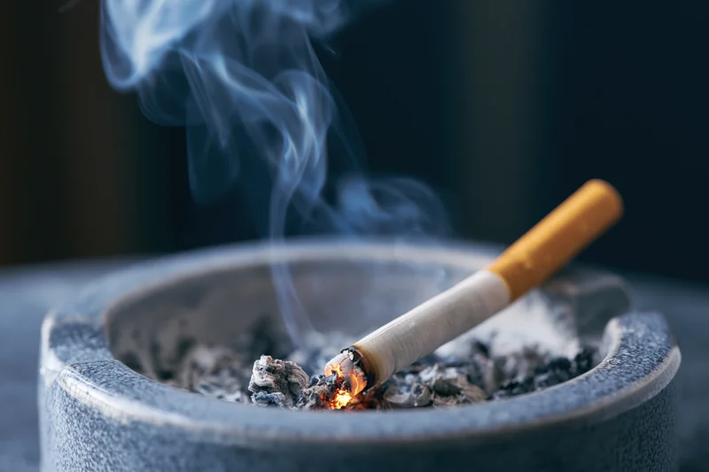 Lit cigarette burning in an ash-filled ceramic ashtray with rising smoke