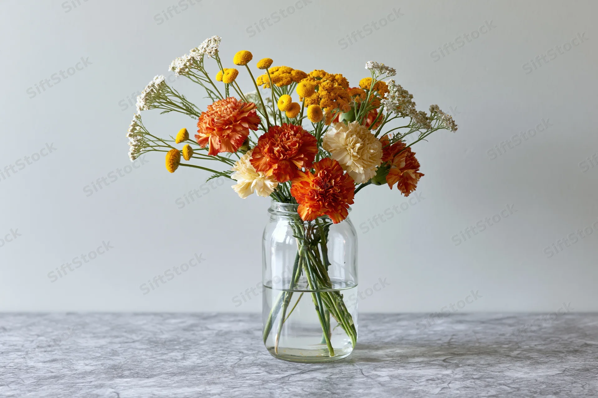 Mixed bouquet of orange carnations, yellow marigolds, and white yarrow in a glass vase on a gray surface