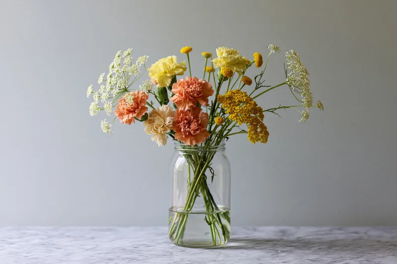 Mixed bouquet of carnations, dill, and tansy in a glass mason jar on a marble table