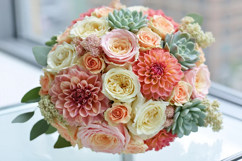 Lush bridal bouquet of garden roses and dahlias in coral and peach tones with succulents on a glass surface near a window