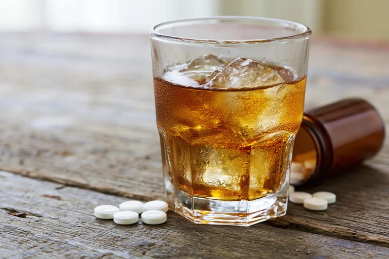 Whiskey in a rocks glass with ice and scattered pills on a wooden table